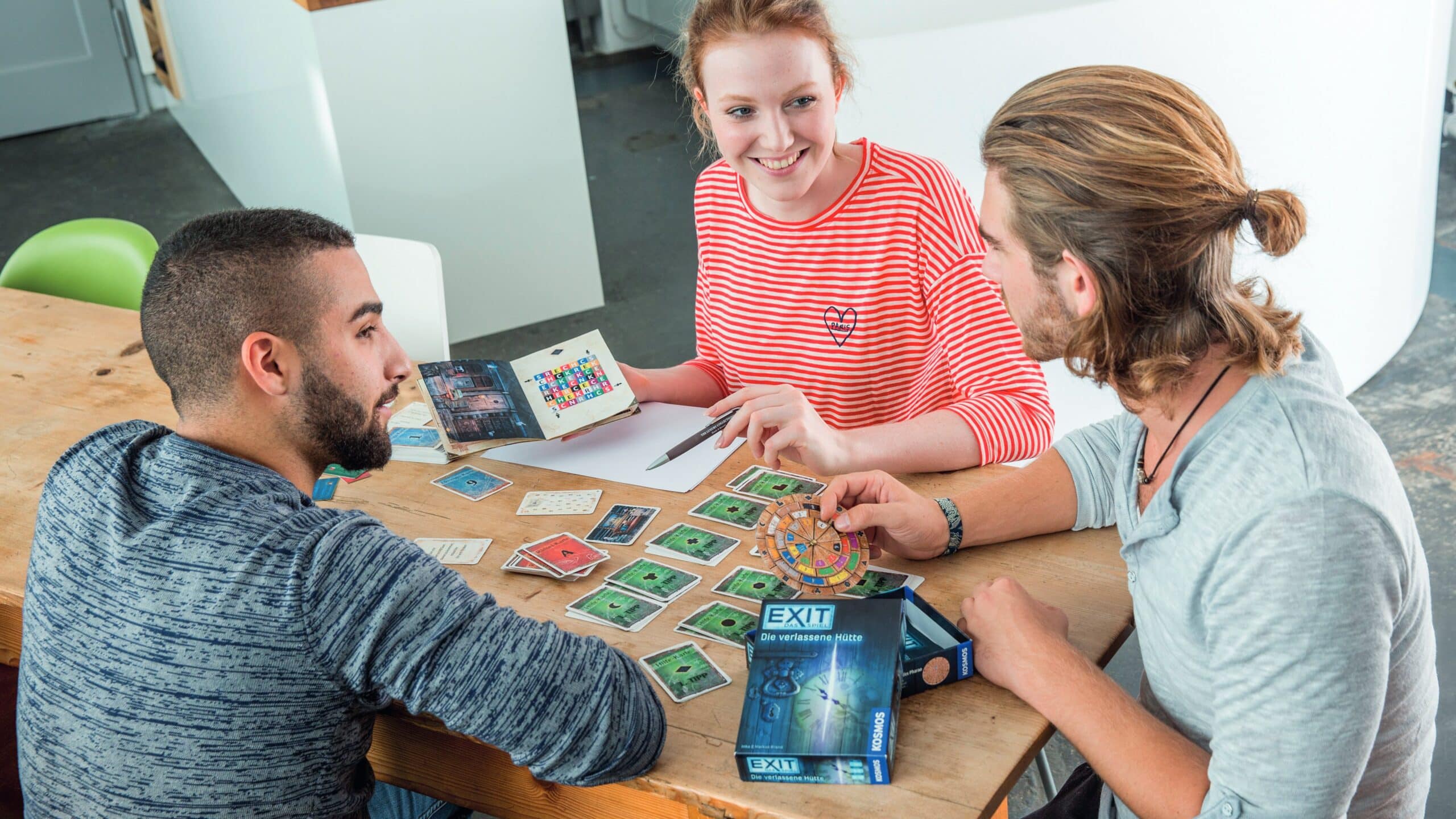 Drie vrienden zitten aan een tafel en spelen het EXIT-spel The Abandoned Hut.